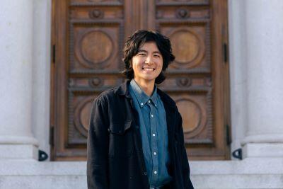 Daniel Lee wearing a black jacket and blue denim shirt standing in front of decorative wood door
