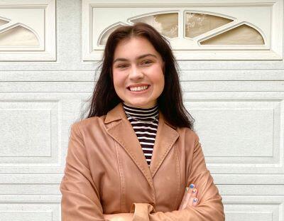 Kylee Wiens wearing brown jacket with black and white striped shirt standing in front of white garage door
