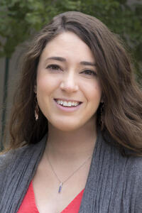 Ashley Noel Metzger headshot wearing gray and red tops with green tree in background.