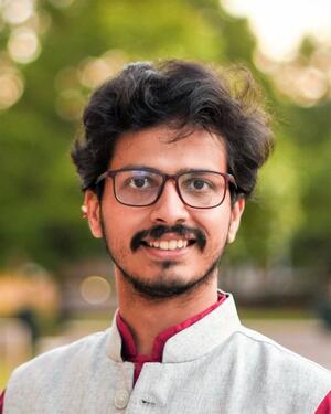 Aakash Chowkase using glassess with gray vest and red shirt headshot with greenery background
