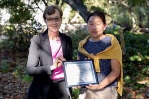 Vice Chancellor for Research Dr. Katherine Yelick presents the faculty postdoc mentor award to Professor Ting Xu with trees and shrubbery in background. Both are holding the certificate.