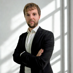 Postdoc Marco Maurizio standing with arms crossed wearing a black jacket and white shirt with white wall and shadows in background