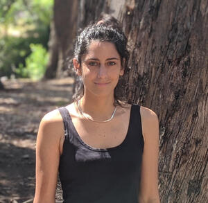 Postdoc Mora Ogando standing in front of a tree with greenery in background.