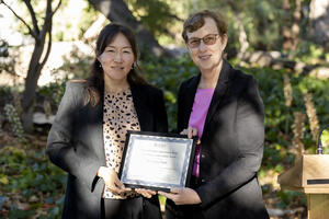 Dr. Lenore Pipes is presented the Postdoc Achievement Award from Vice Chancellor for Research Dr. Katherine Yelick with trees and shrubbery in background. Both are holding the certificate.