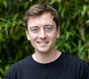 Tomas Hessler wearing a black tshirt standing in front of green shrubbery in the background