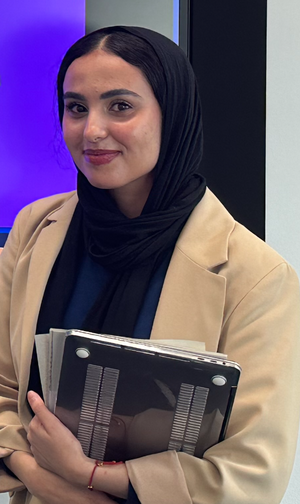 Nouf Ali S AlGazlan wearing a black top and a tan coat holding a laptop and books